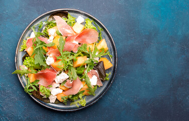 Summer salad with cantaloupe melon, prosciutto, white cheese and arugula on blue table background, top view, copy space