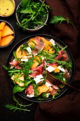 Summer salad with melon, jamon, white cheese and arugula on brown table background, top view, copy space