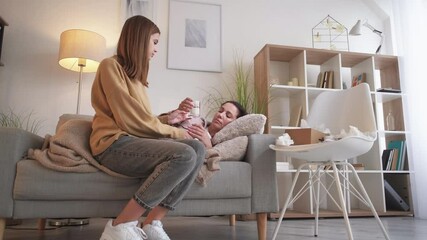 Family care. Fever mother. Virus infection. Careful teenager daughter bringing glass of water to sick woman laying under plaid on sofa in light room interior.