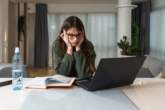 Young freelancer woman working at home sitting at the kitchen countertop loosing her concentration because of slow internet connection. Female worker having issue with old laptop computer.