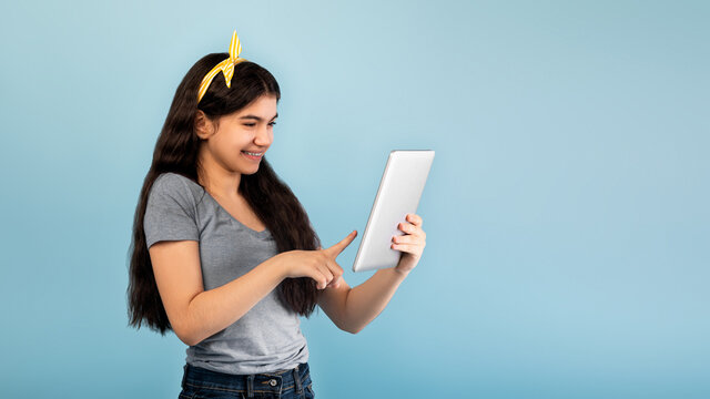 Portrait of Indian teen girl using tablet computer, studying online on blue studio background, panorama with copy space