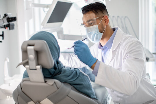 Middle-Eastern Male Dentist In Medical Mask And Face Shield Checking Patient's Teeth