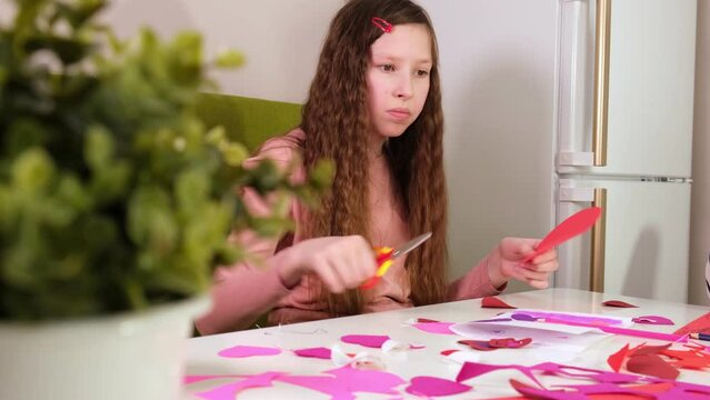Little Girl with paper heart. Children, doing Valentine's Day arts and crafts with scissors, pencils. Kids Crafts Heart, tracing and cutting out from pink paper for Valentines Day. Painting, DIY