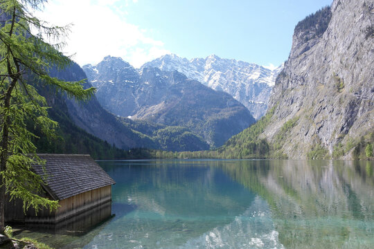 Wooden boat house, serene Alps snowy peaks landscape. Calm clean azure water surface. Inspiring fresh mountain air. Natural time of clarity of thoughts.  Sunny Obersee lake shore, Bavaria, Germany. 