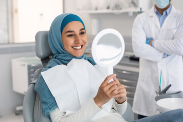 Happy Muslim Female Patient Looking At Mirror After Treatment In Dental Clinic