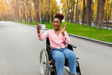 Happy young black disabled lady in wheelchair having video call on smartphone, waving at camera in...