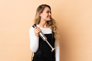 Young brazilian woman using hand blender isolated on beige background looking side