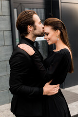Portrait of charming man gently kissing gorgeous smiling woman, having a lovely date. Beautiful happy couple in fashion black outfits walking at the city, hugging, enjoying moments together
