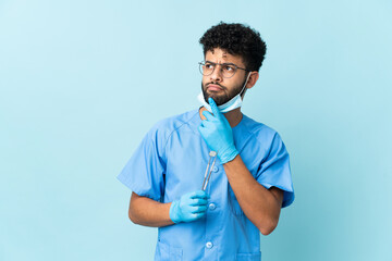 Moroccan dentist man holding tools isolated on blue background having doubts while looking up