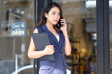 Portrait of beautiful Asian casual businesswoman talking by smartphone while standing in front cafe. Young entrepreneur making business calls by a mobile phone and holding laptop while go to office.