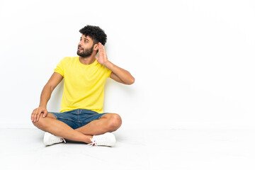 Fototapeta premium Young Moroccan handsome man sitting on the floor over isolated background listening to something by putting hand on the ear