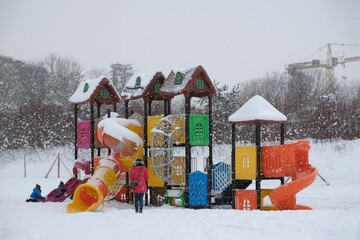 Colorful playground for children on beach on winter day. Children are playing, mother is standing by them. It's snowing heavily.