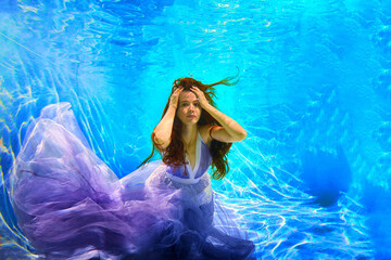 Girl in a beautiful dress under blue water. Female model posing under water in swimming pool