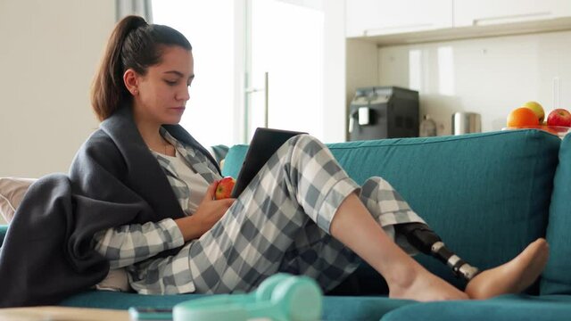 Handsome woman with artificial bionic high technology leg prosthesis texting by tablet and eating apple at home