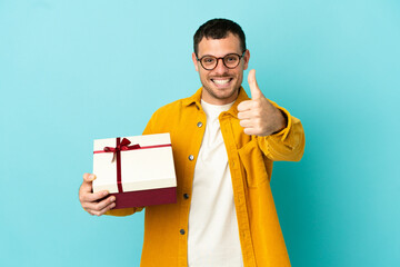 Brazilian man holding a gift over isolated blue background with thumbs up because something good...