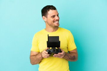 Brazilian man holding a drone remote control over isolated blue background looking to the side and smiling