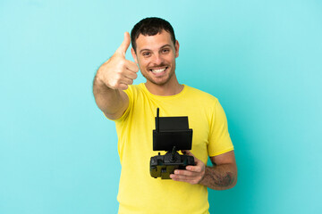 Fototapeta na wymiar Brazilian man holding a drone remote control over isolated blue background with thumbs up because something good has happened
