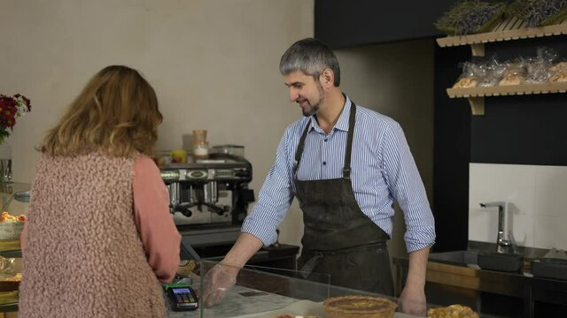 Handsome male cashier in apron and gloves accepts terminal payment from female customer