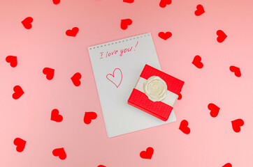 A white sheet of a notebook with the inscription "I love you" and a beautiful gift box on a pink background with small red hearts. Top view, festive, creative composition. Concept - surprises and gift