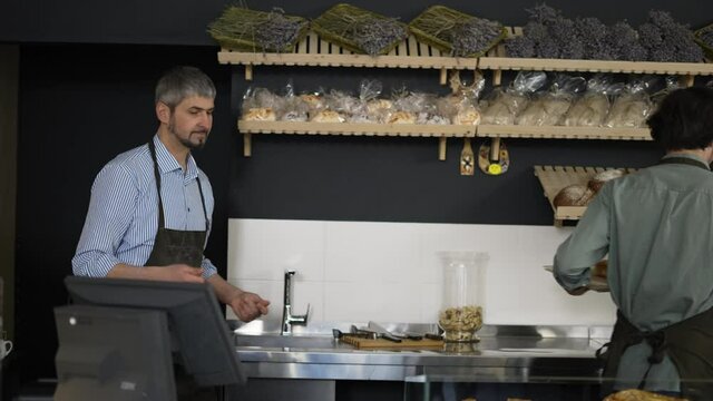 Handsome father in apron passes a tray of pastries to his young son and enters data into the bakery database. Indoor, close up, slow motion