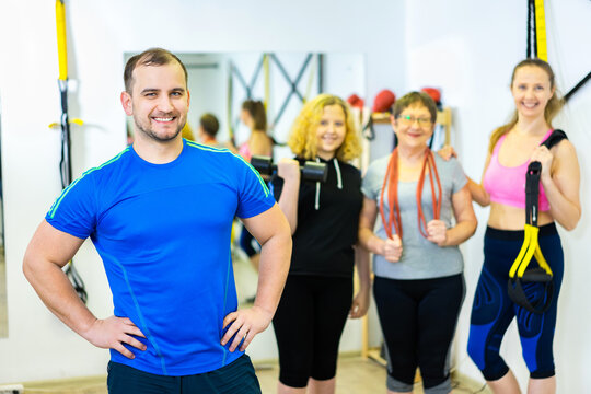 In The Foreground, A Cheerful Male Trainer Smiles And Rests His Hands On His Hips. Happy Sports Family Behind Man. Grandma, Mom, Daughter.