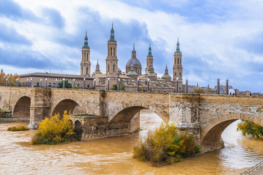 Zaragoza landscape with ancient Stone Bridge over Ebro river with yellow water and Cathedral-Basilica of Our Lady of the Pillar in the background. Panorama of historical spanish town in autumn day
