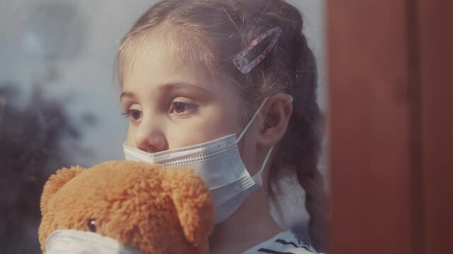 sad kid girl in protective medical mask by the window looks out. stay home coronavirus teddy bear concept. sad kid in protective mask in hospital looking out window. stay home coronavirus pandemic