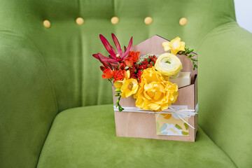 Giftbox with flowers and greeting card. Colorful spring bouquet in wooden box on green soft armchair.