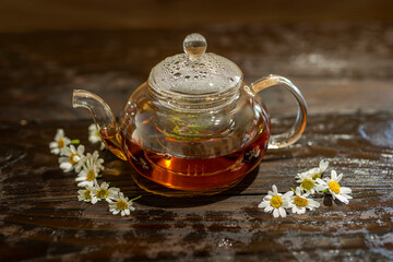 Steaming glass teapot with chamomile tea on a wet wooden table.