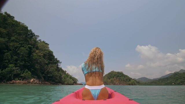 Sexy young woman in blue swimsuit swings feet in water on pink canoe on sea against hills backside view.