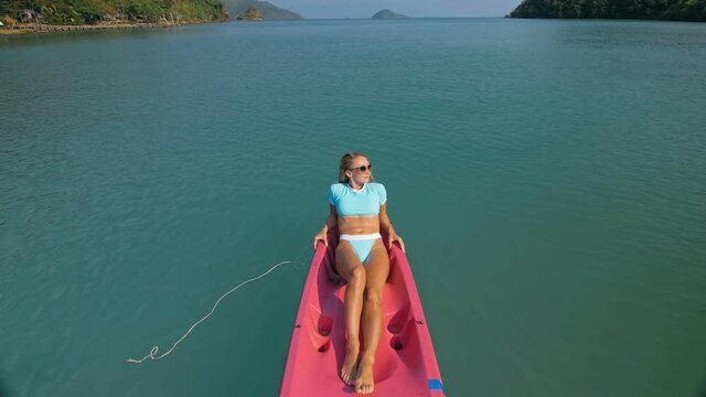 Pretty slim woman in elegant swimsuit lies in pink plastic kayak drifting on azure ocean at exotic resort upper view.