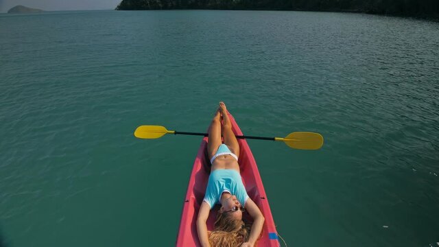 Pretty slim woman in elegant swimsuit lies in pink plastic kayak drifting on azure ocean at exotic resort upper view.