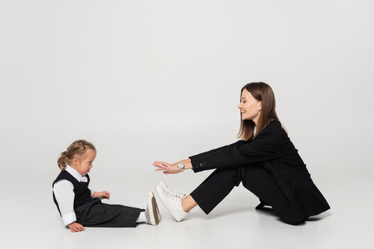 Side View Of Cheerful Mother Sitting And Reaching Child With Down Syndrome On Grey.