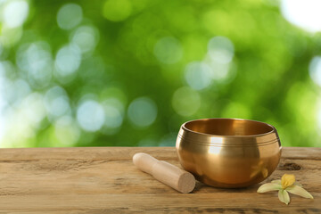 Golden singing bowl, mallet and flower on wooden table against outdoors, space for text