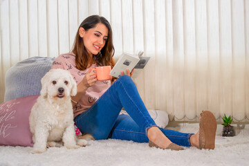 Beautiful young Latin woman with brown hair and dressed in pink, sitting on a white fluffy rug with cushions by the window, enjoying a moment in the living room with a book and her puppy dog.