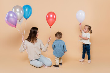 cheerful mother sitting near girl with down syndrome and toddler boy holding colorful balloons on...