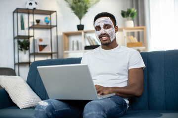 Indoor portrait of handsome African American man with white clay mask on face, sitting on couch at home and working on laptop and having hygiene skin care procedures