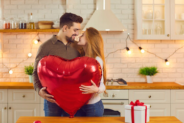 Cute couple standing in kitchen, holding huge shiny red heart-shaped balloon and kissing. Happy young woman enjoying romantic presents and surprises from boyfriend on anniversary or Valentine's Day