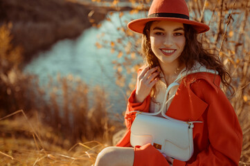 Happy smiling fashionable woman wearing trendy orange color hat, autumn trench coat, with white faux leather bag, posing in nature. Copy, empty space for text