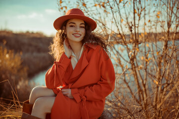 Happy smiling fashionable woman wearing trendy orange color hat, autumn trench coat, posing in nature. Copy, empty space for text