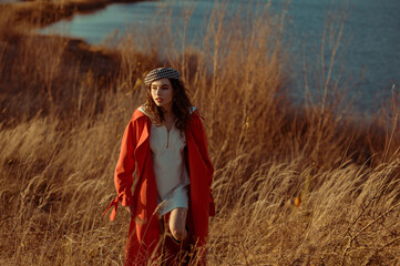 Elegant fashionable woman wearing trendy outfit: houndstooth printed beret, orange trench coat, white mini dress, walking in nature. Outdoor autumn portrait. Copy, empty space for text