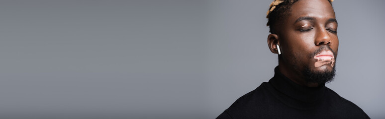 young african american man with vitiligo and earphone listening music with closed eyes isolated on grey, banner.