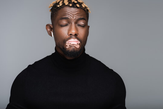 african american man with vitiligo standing with closed eyes while listening music in earphone isolated on grey.