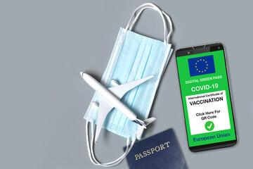 Smartphone with european digital covid-19 vaccination certificate along with protective masks, airplane model and passport on grey background. Safe travel concept