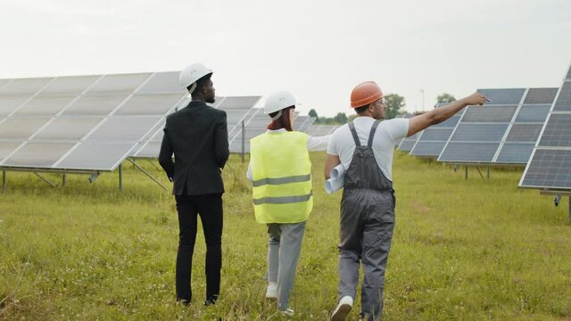 Back view of two inspectors in white helmets walking on solar station with technician in uniform. Multiracial engineers examining work of photovoltaic cells outdoors. Workers walking on solar station.