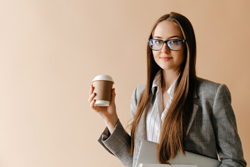 Young confident businesswoman posing with coffee cup, holding laptop and papers isolated on light brown background. Time is business concept