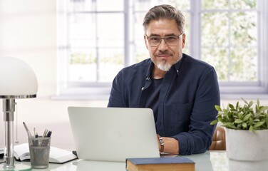 Casual businessman working with laptop computer in home office, happy, smiling. Portrait of middle age man in 50s, white caucasian, gray hair. - obrazy, fototapety, plakaty