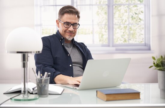 Casual businessman working with laptop computer in home office, happy, smiling. Portrait of middle age man in 50s, white caucasian, gray hair.