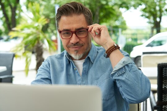 Outdoor portrait of mid adult man in 50s, happy confident smile. Working with laptop computer. Mature age, middle age, bearded, glasses.
