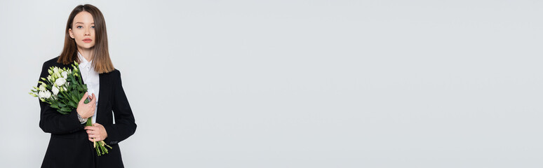 trendy young woman in black suit holding flowers isolated on grey, banner.